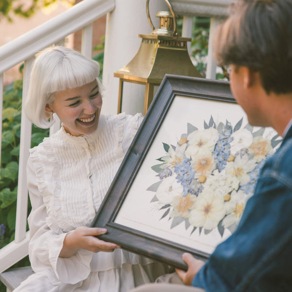 Woman in white dress holding a framed floral artwork on a porch with a man.