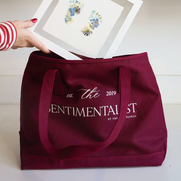 Person holding a framed print above a maroon tote bag with 'The Sentimentalist' branding.