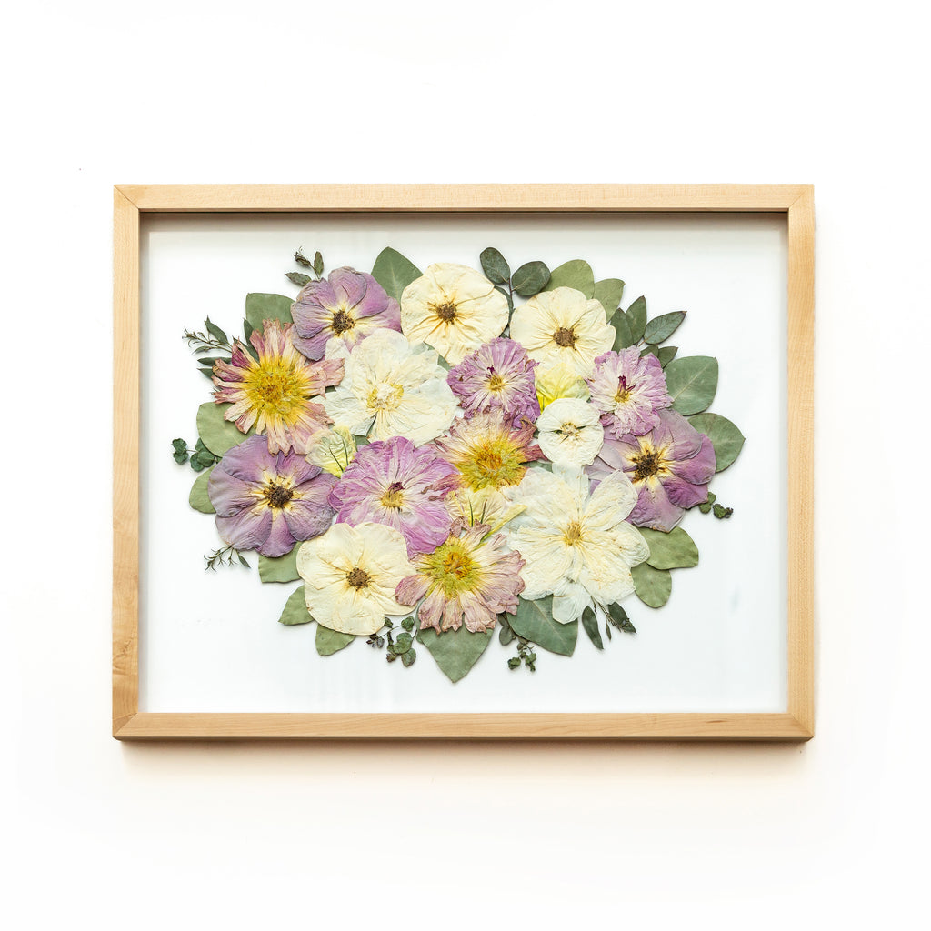 Framed pressed flower art featuring a vibrant arrangement of white, purple, and yellow flowers with green leaves, set against a white background.