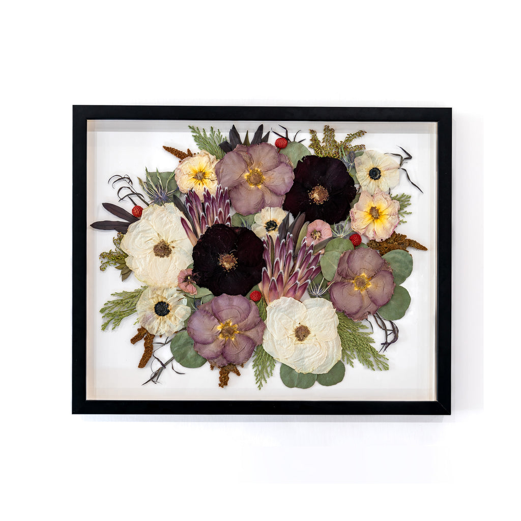 A sophisticated 16x20 sleek black frame highlighting a captivating display of pressed flowers. The arrangement includes elegant purple roses, pristine white roses, and striking proteas, expertly preserved to capture their timeless allure.