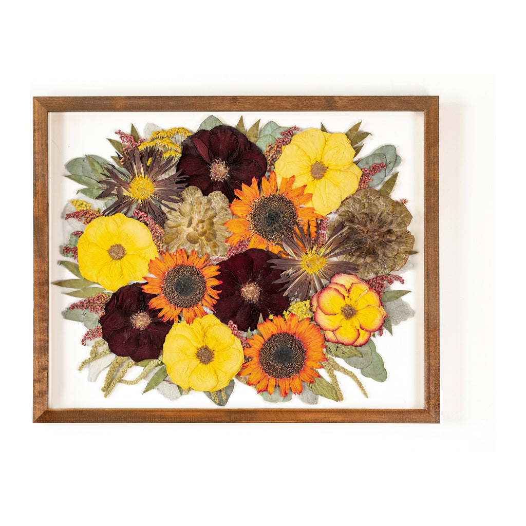 A walnut brown frame showcasing a vibrant arrangement of preserved flowers. Sunflowers, with their bold yellow petals and dark centers, stand tall amidst a backdrop of lush succulents. Interspersed among them are sunny yellow roses, adding a delicate touch to the composition. The warm tones of the walnut brown frame beautifully complement the cheerful colors of the preserved flowers, creating a visually striking display that exudes energy and natural beauty.