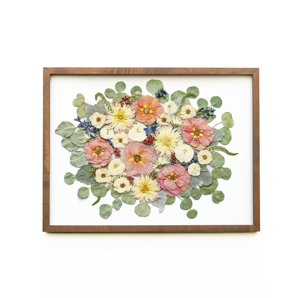 A walnut brown frame beautifully adorned with preserved flowers in soft hues. Delicate pink and pristine white roses intermingle with elegant white dahlias, forming an exquisite composition. The warm tones of the walnut brown frame provide a lovely contrast, highlighting the gentle colors of the preserved flowers. This arrangement emanates a sense of grace and purity, creating a visually captivating display that is both timeless and elegant.