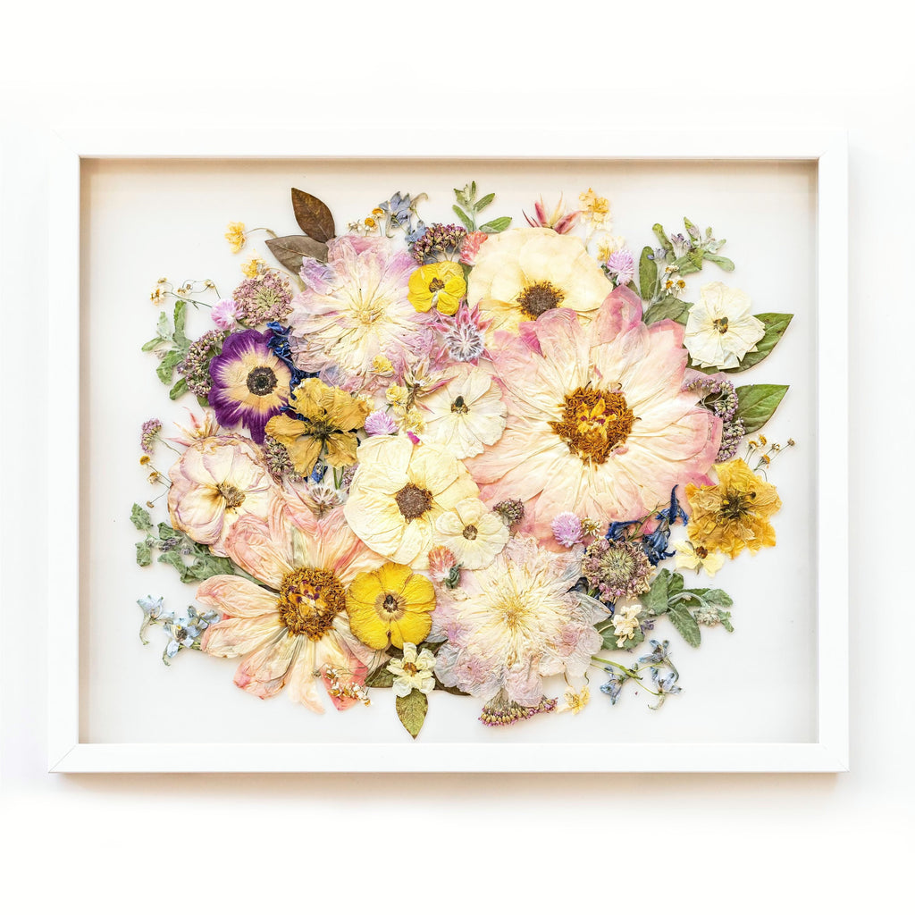 A white frame showcasing a captivating arrangement of preserved flowers. Soft pink peonies, with their lush and voluminous petals, bloom alongside delicate pink anemones and vibrant yellow ranunculus. The combination of these flowers creates a harmonious blend of colors and textures. Against the pristine white frame, the preserved flowers exude a sense of elegance and grace. This composition evokes a feeling of joy and vibrancy, capturing the beauty of nature in a delightful display.