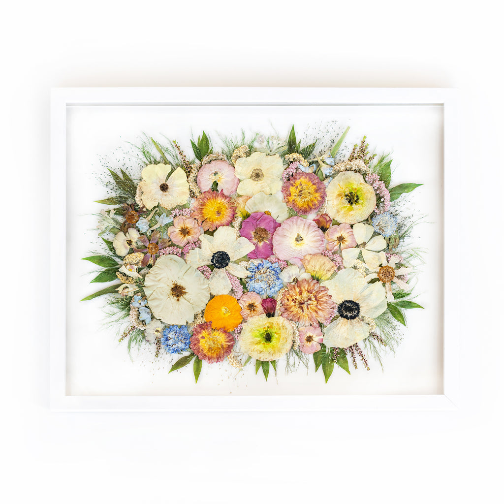 A charming 16x20 solid white frame showcasing a delightful arrangement of pressed flowers. The composition features delicate anemones, graceful pink dahlias, pristine white roses, and enchanting pink ranunculi, artfully preserved to capture their captivating beauty.